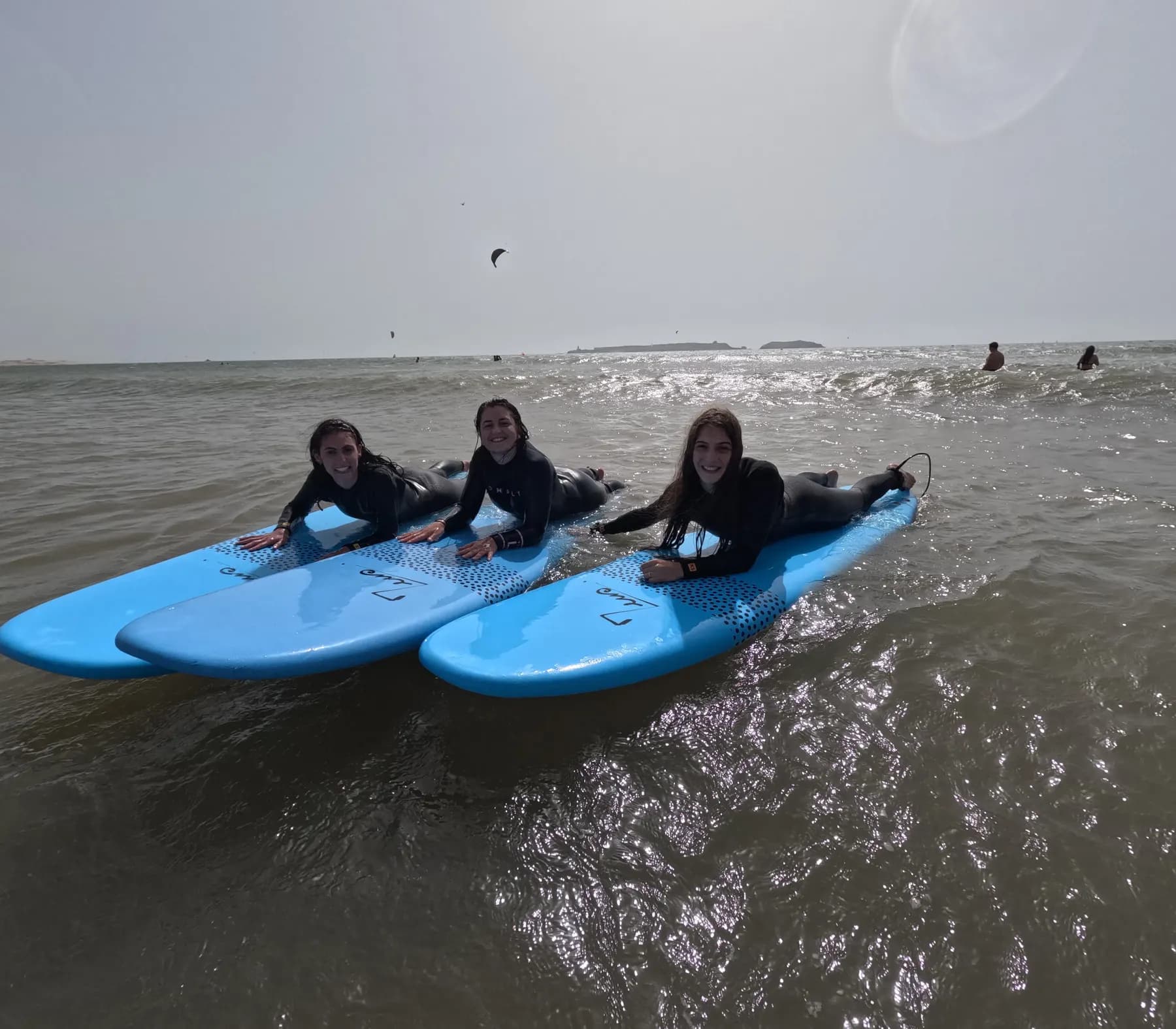 Three young surfers in wetsuits lying on light-blue foam boards in shallow water, with low islands on the horizon